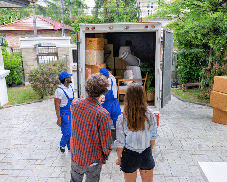 Movers are unloading boxes and furniture from a truck outside a house, while two people observe the process. The scene conveys a busy and organized moving day.