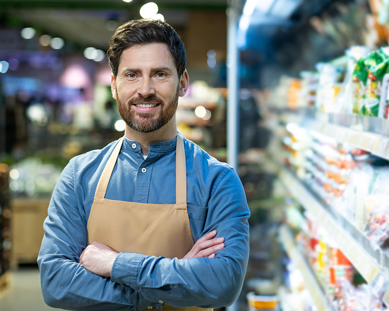 A smiling man wearing an apron stands confidently with his arms crossed in a grocery store aisle, surrounded by various products on the shelves.