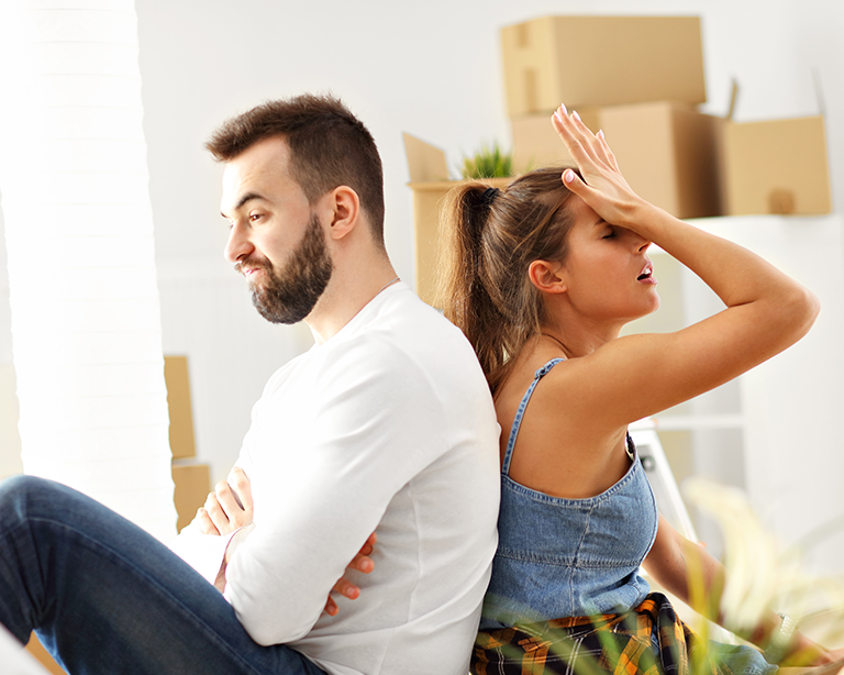 A couple sits back-to-back with a frustrated expression, surrounded by moving boxes, conveying the stress often associated with relocation.