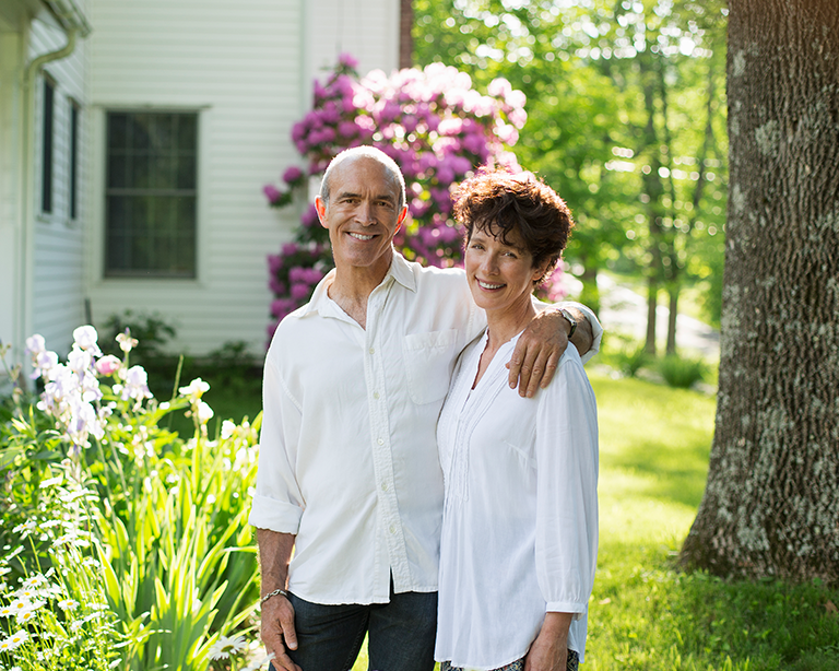 A couple stands together in a lush garden, smiling warmly at the camera, surrounded by vibrant flowers and greenery. The scene conveys a sense of warmth and partnership.
