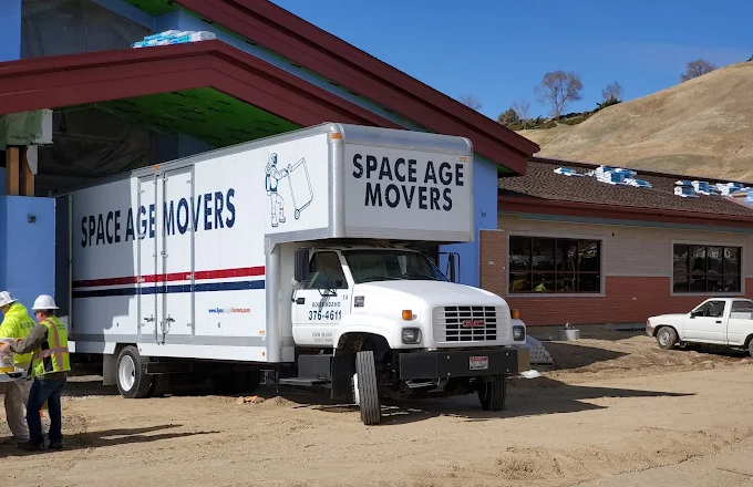 A moving truck labeled "Space Age Movers" is parked outside a building under construction, with workers in safety gear nearby. The scene captures the essence of a reliable moving service in action.