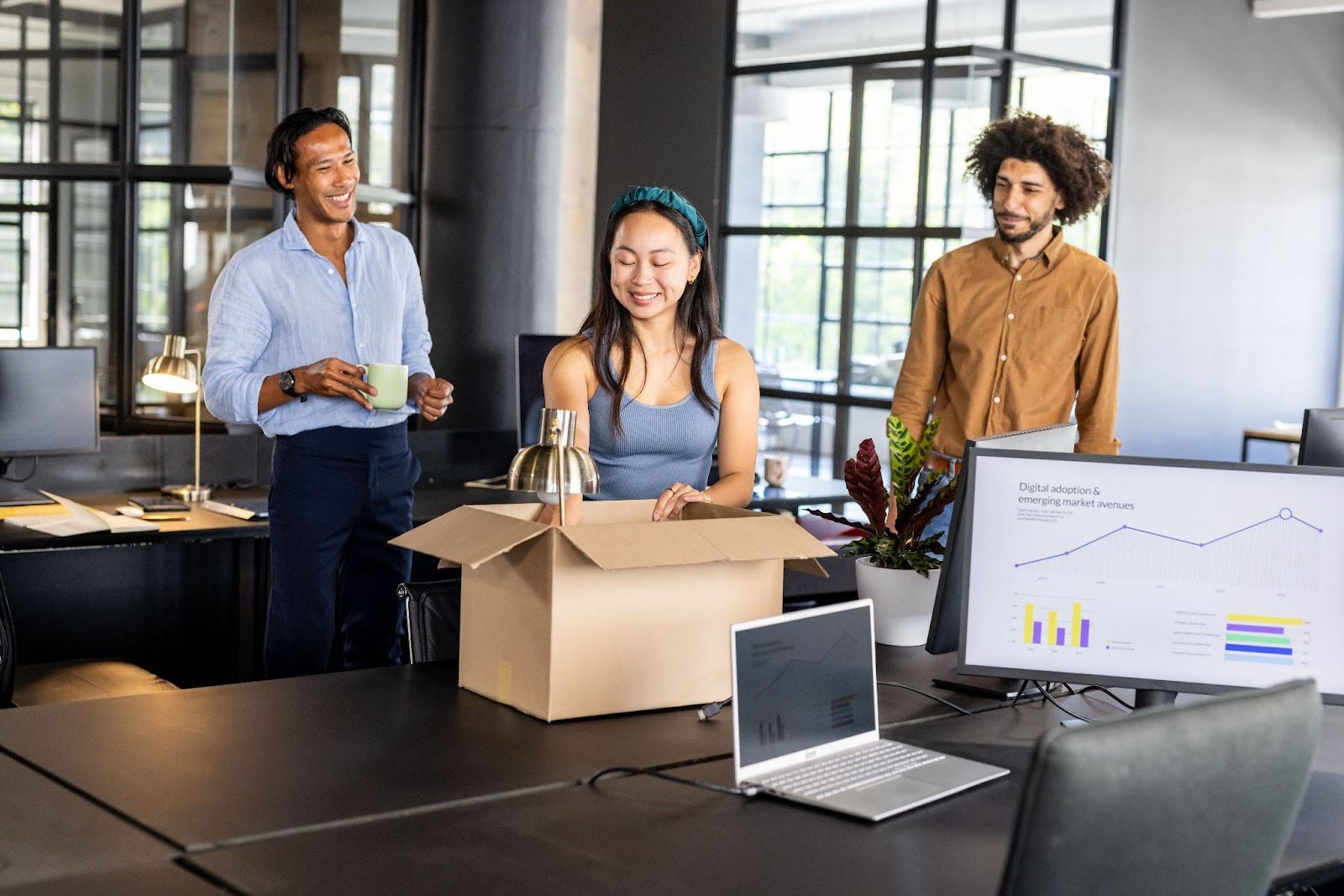Three colleagues are gathered around a table in a modern office, with one person unpacking a box while the others watch and smile. A laptop and charts are visible on the table, suggesting a collaborative work environment.
