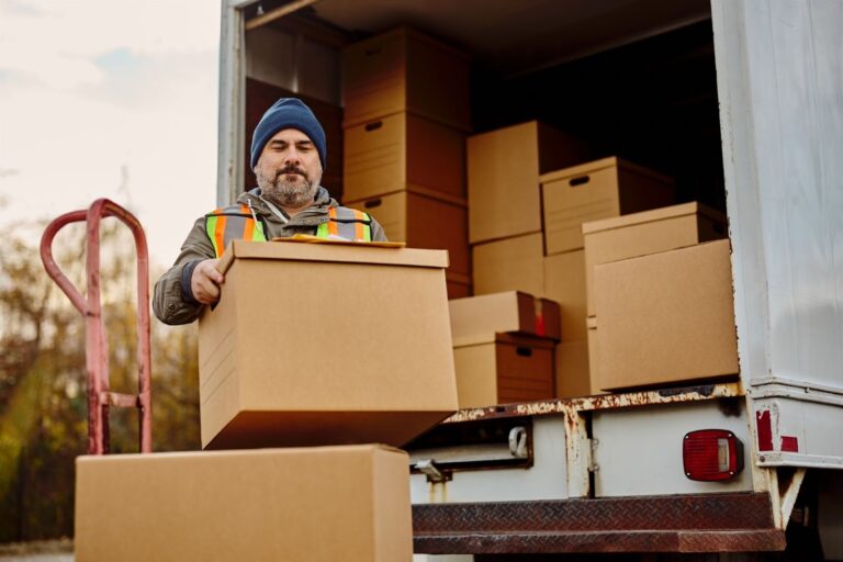 A worker in a beanie and reflective vest is loading a cardboard box from a moving truck, surrounded by stacked boxes. The scene captures the process of moving and relocation services.