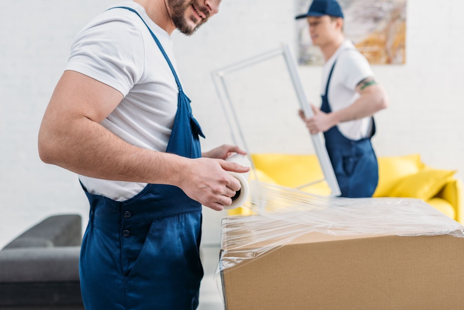 Two movers are packing a large cardboard box in a bright living room, with one applying tape while the other holds a picture frame.