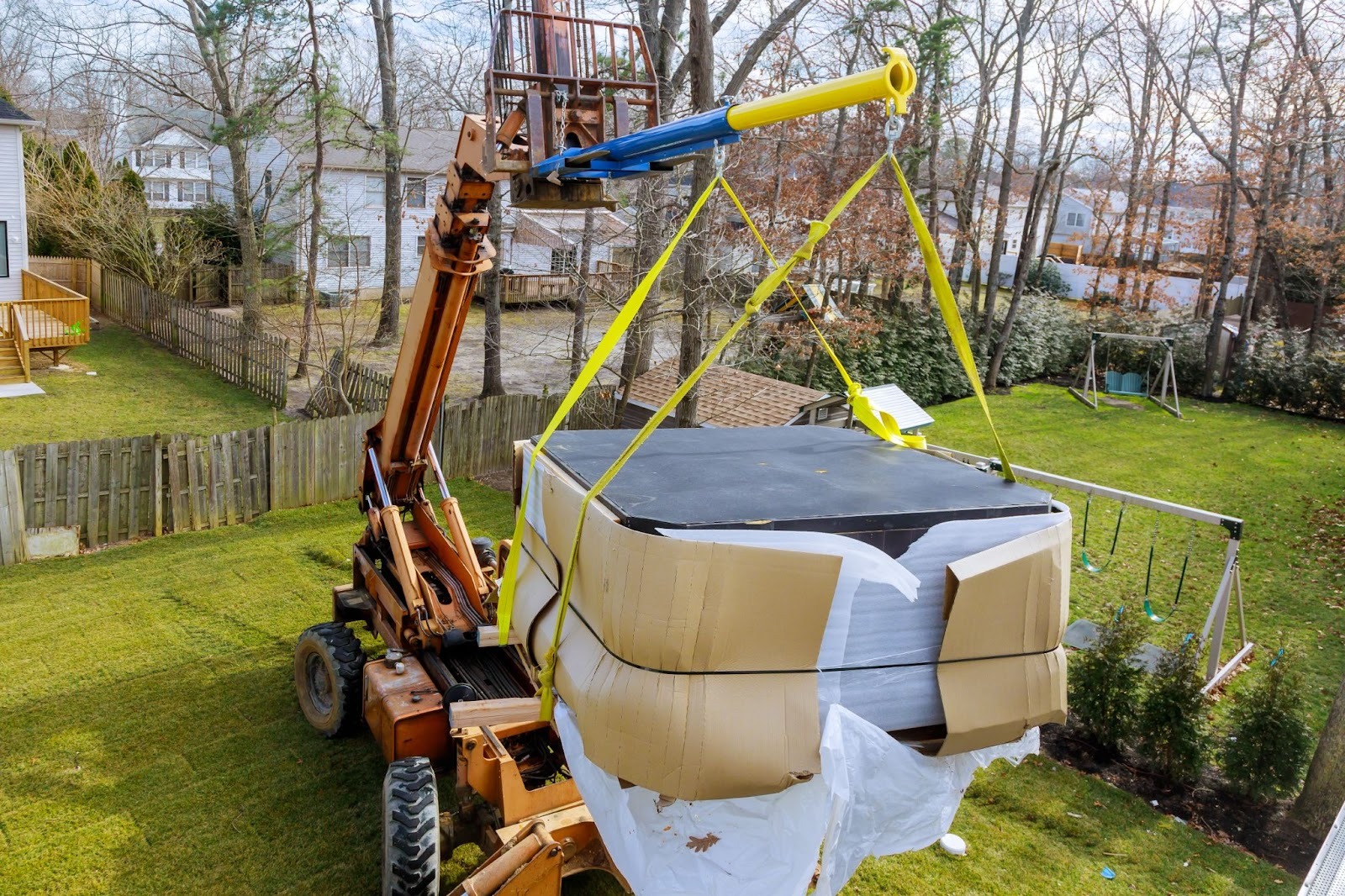A forklift carefully hoists a large, wrapped hot tub over a green yard, surrounded by trees and a wooden fence on a clear day. Residential scene.