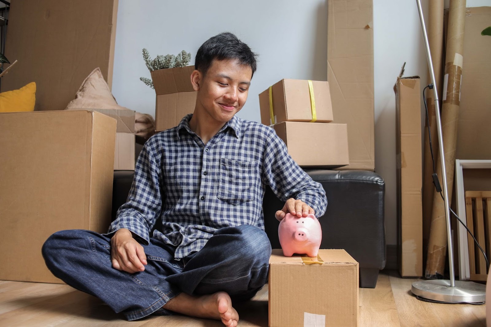 A young man sits on the floor surrounded by cardboard boxes, smiling as he places a coin into a pink piggy bank.