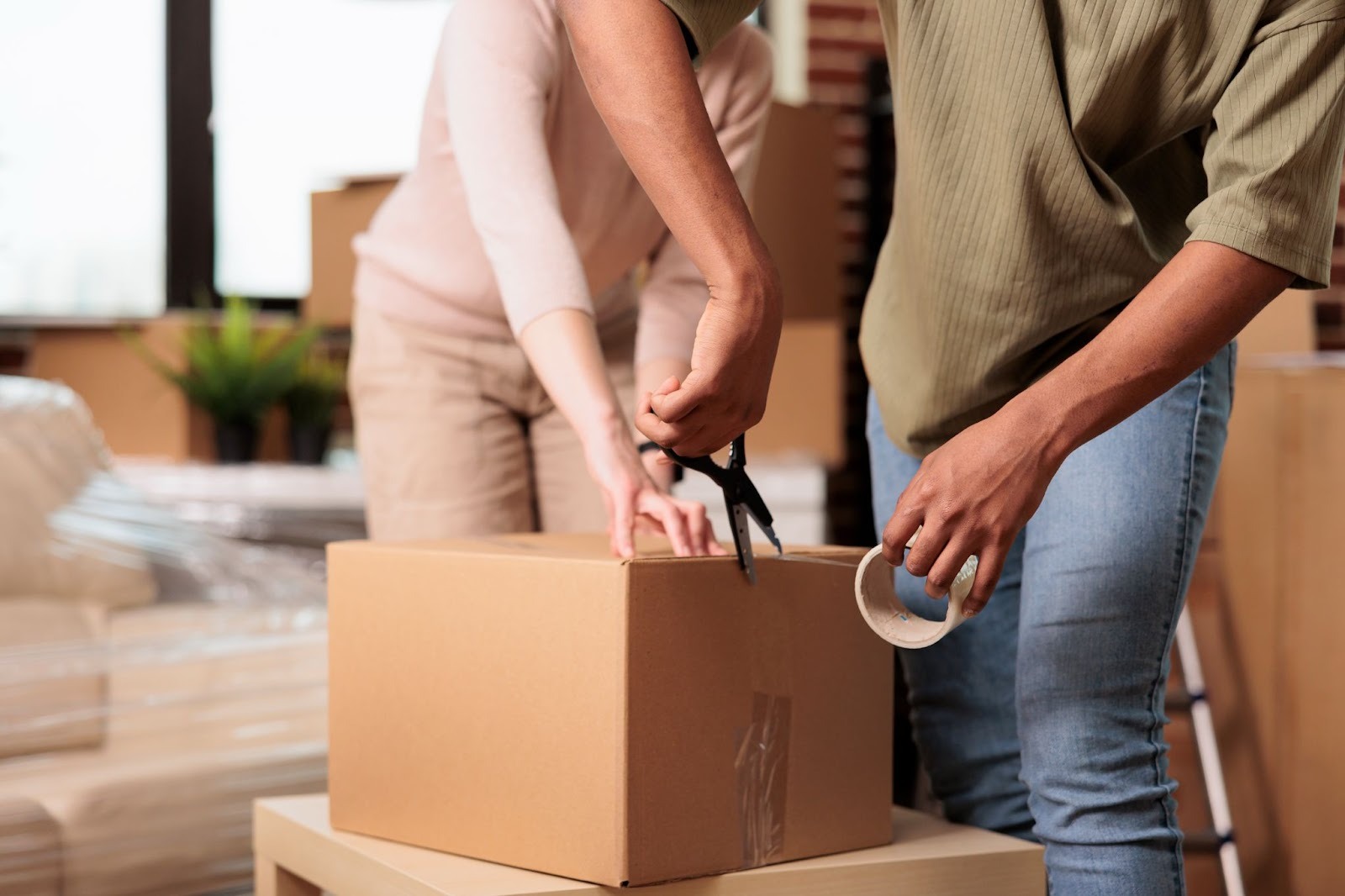 Two people are preparing a cardboard box for packing, one is using scissors while the other holds a roll of tape. The setting appears to be a room with moving supplies.