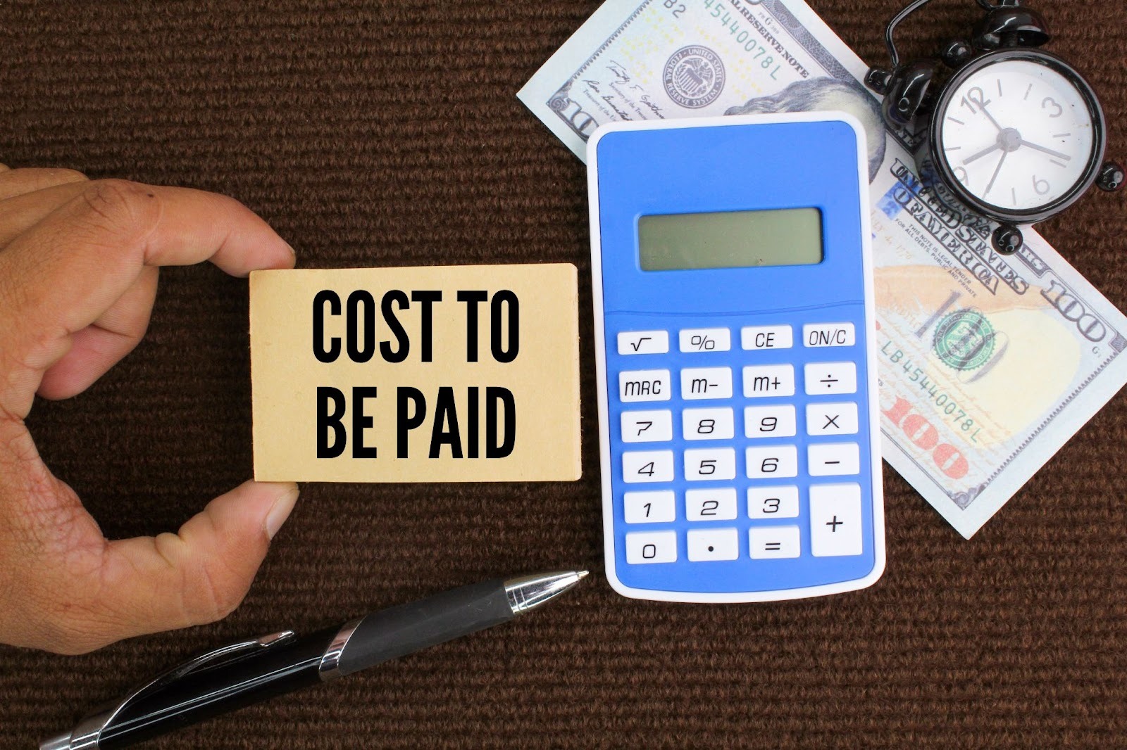 A hand holds a card reading "COST TO BE PAID" next to a blue calculator, pen, cash, and an alarm clock on a brown surface, conveying a theme of financial urgency.