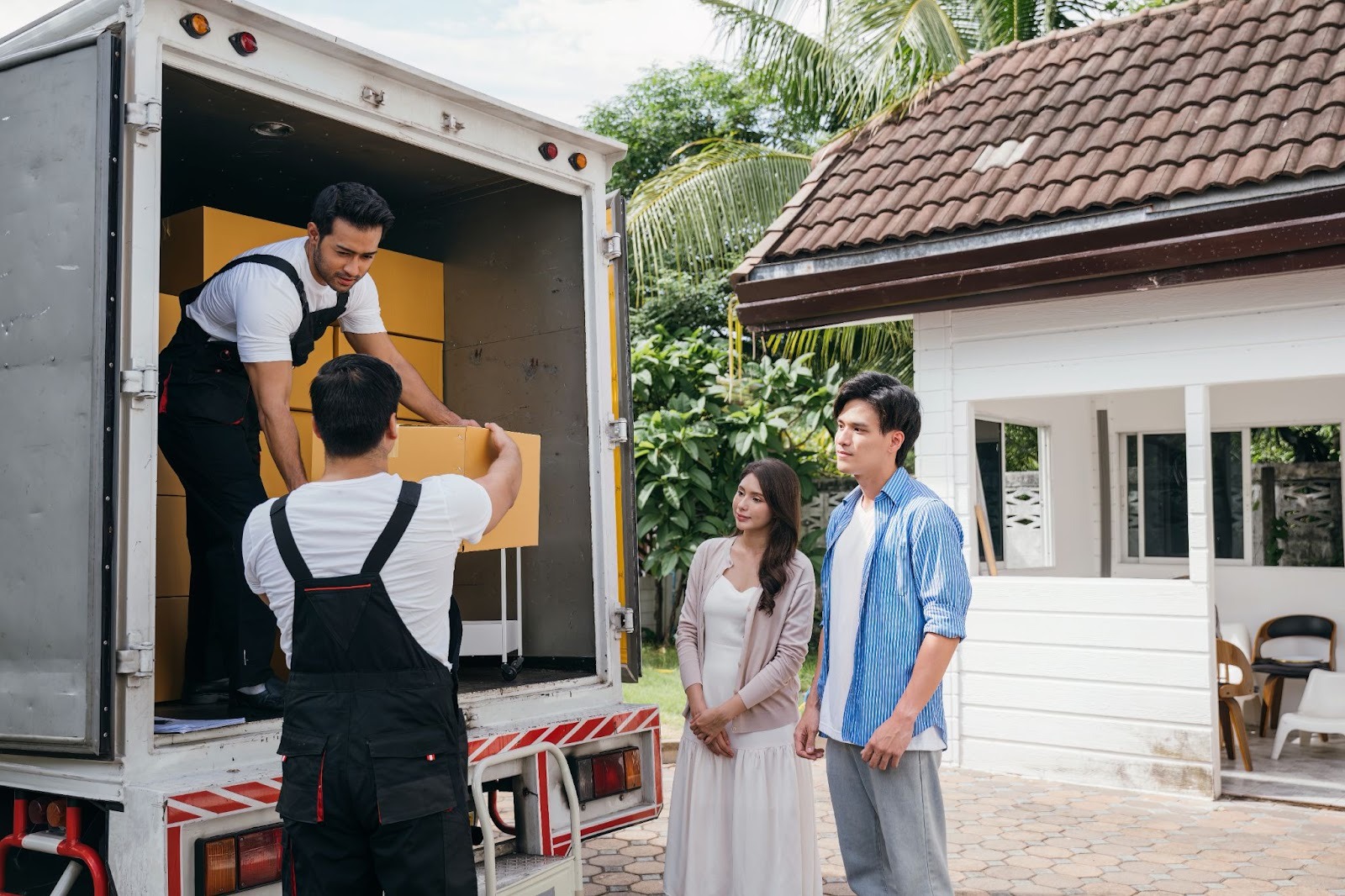 Two people stand outside a house while a third person unloads boxes from a moving truck. The scene captures a moment of moving into a new home, set in a suburban environment.