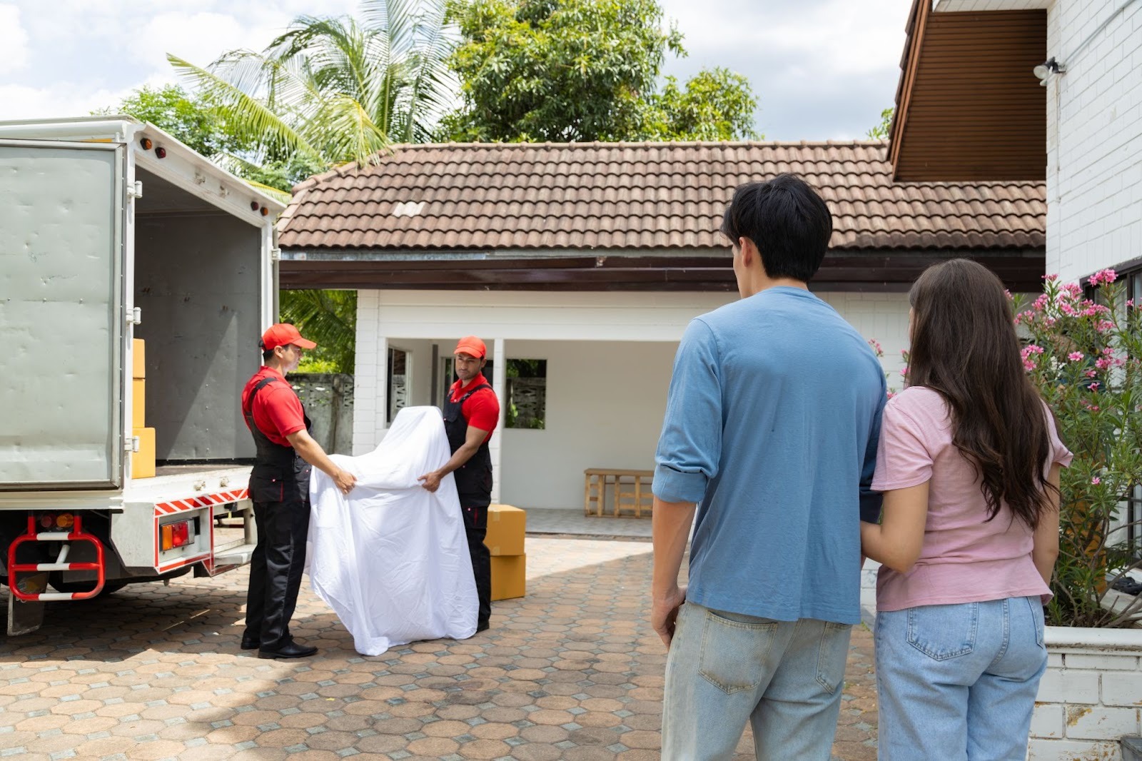 Two movers in red uniforms unload furniture from a truck into a driveway, observed by a couple standing nearby. The scene conveys a sense of anticipation.