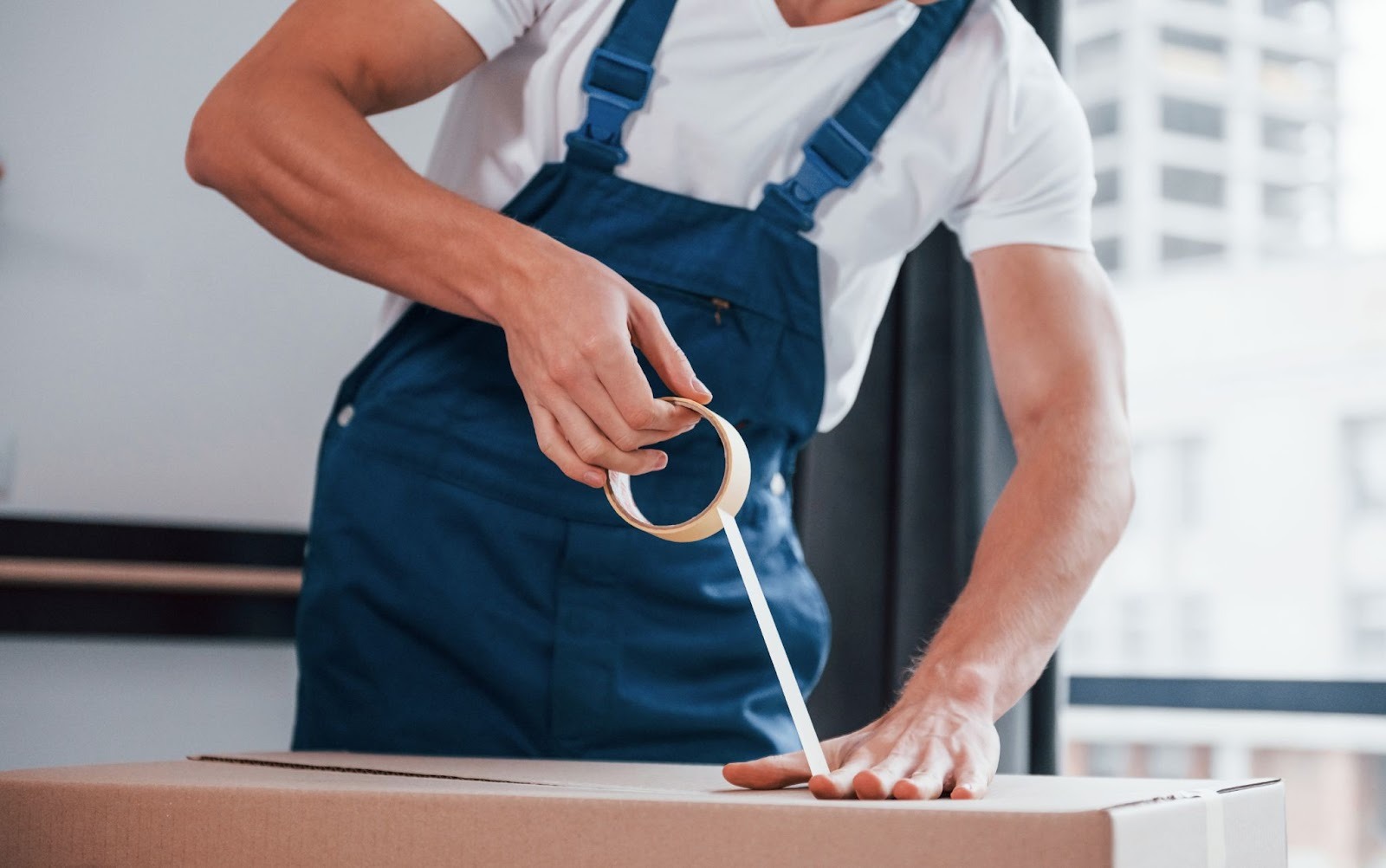 A person in blue overalls is sealing a cardboard box with packing tape in a bright indoor space.
