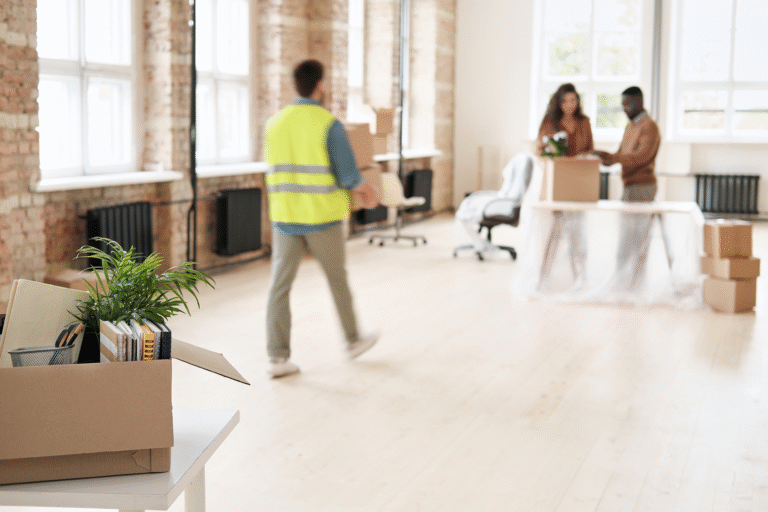A person in a safety vest walks past a couple discussing office items in a bright, spacious room filled with moving boxes and plants. The setting suggests a busy office relocation process.