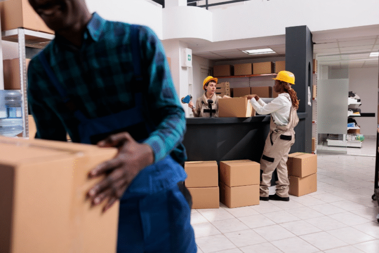 A group of movers is working in a spacious indoor setting, handling cardboard boxes and coordinating their efforts. The scene conveys a sense of teamwork and efficiency in the moving process.