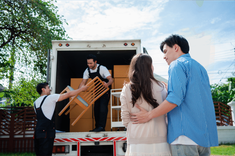 A couple watches as movers unload furniture from a moving truck in a residential area, showcasing a seamless relocation process. The scene conveys a sense of teamwork and support during the moving experience.