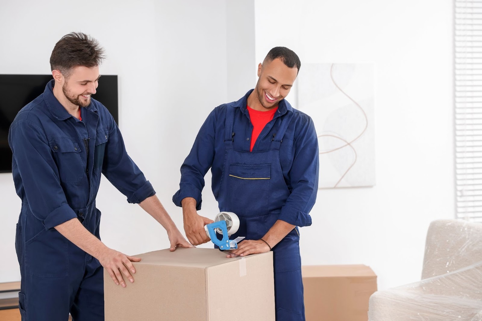 Two professional movers sealing a moving box with packing tape inside a home during relocation