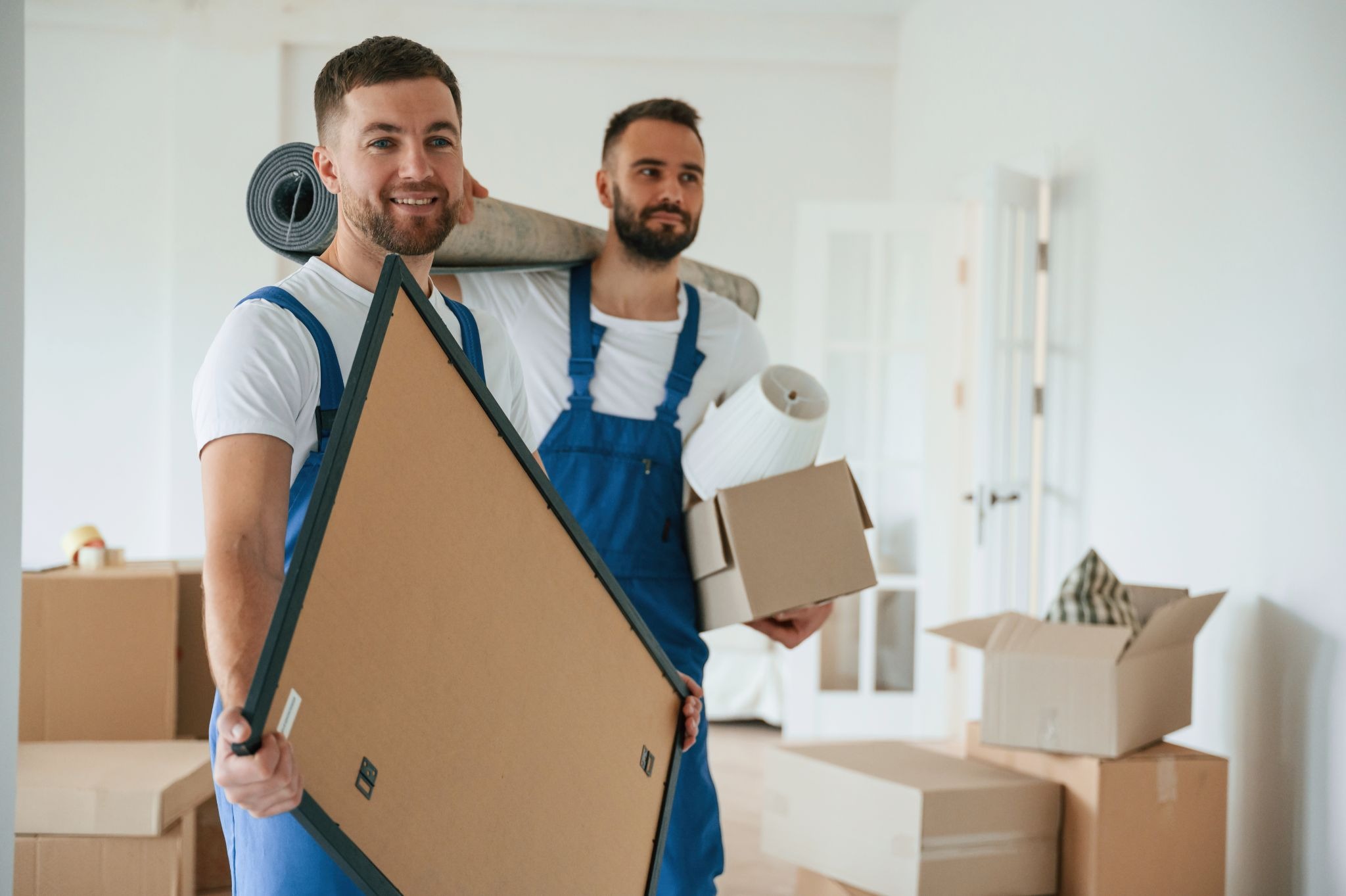 Two professional movers in white shirts and denim overalls carry boxes into a new home